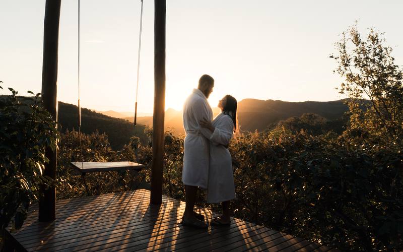 Casal em roupões no deck do balanço da Villa Eyá com o sol se pondo sobre o Vale do Moinho